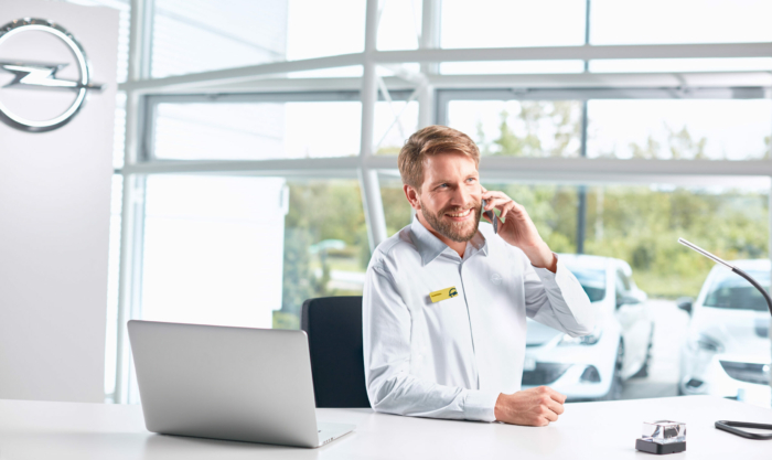 Ein Opel-Mitarbeiter sitzt in einem modernen Autohausbüro, telefoniert freundlich mit einem Kunden und arbeitet am Laptop.