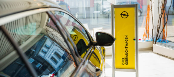 Schaufenster eines Autohauses mit gelbem Aufsteller ‚Opel Gebrauchtwagen‘ und Spiegelung eines Fahrzeugs im Glas.