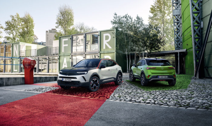 Zwei Opel Mokka Fahrzeuge auf einem farblich gestalteten Platz vor modernen Containerbauten mit großen Buchstaben.