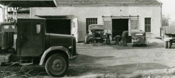Schwarzweißaufnahme einer alten Kfz-Werkstatt mit mehreren Fahrzeugen und arbeitenden Personen vor der Werkstatthalle.