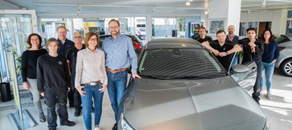 Gruppenfoto des Teams vom Autohaus Burmann in einem hellen Autohaus-Showroom, vor einem silbernen Opel.