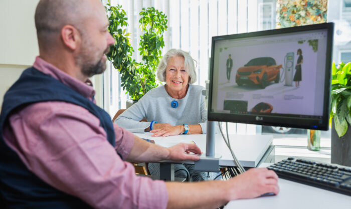 Beratungsgespräch in einem Autohaus: Verkäufer sitzt am Schreibtisch vor einem Monitor, Kundin lächelt im Hintergrund.