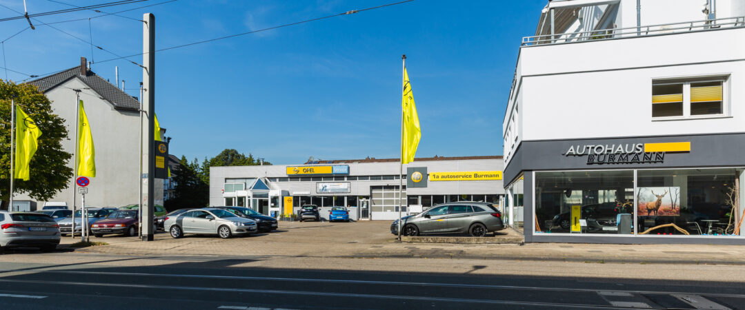 Außenansicht des Autohauses Burmann mit Fahrzeugen auf dem Hof, gelben Opel-Fahnen und dem Firmengebäude bei Tageslicht.