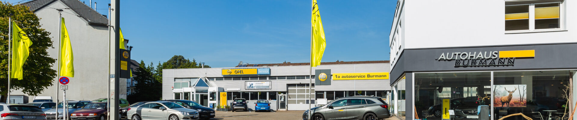 Außenansicht des Autohauses Burmann mit Fahrzeugen auf dem Hof, gelben Opel-Fahnen und dem Firmengebäude bei Tageslicht.