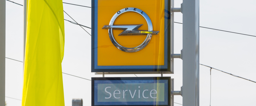 Nahaufnahme eines gelben Opel-Logos mit darunterliegendem Schild ‚Service‘ vor Stromleitungen und Himmelshintergrund.