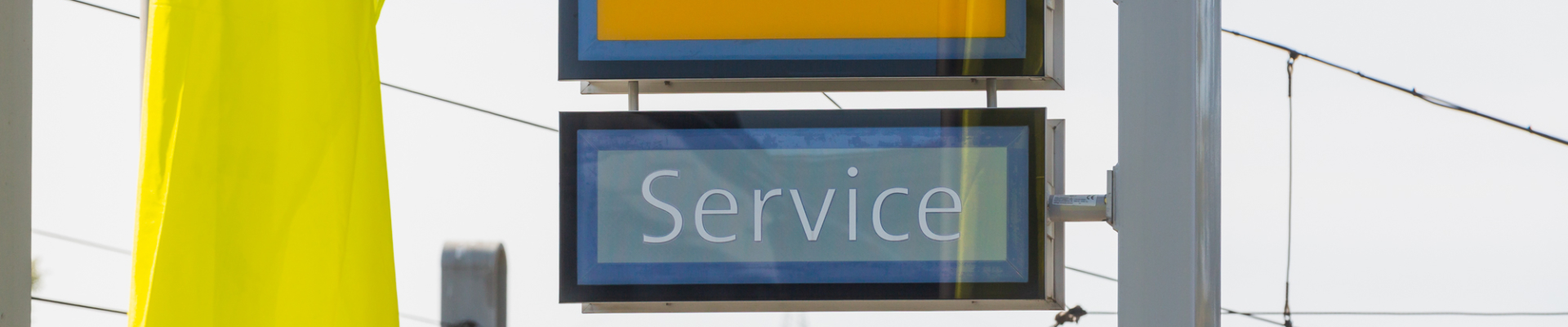 Nahaufnahme eines gelben Opel-Logos mit darunterliegendem Schild ‚Service‘ vor Stromleitungen und Himmelshintergrund.